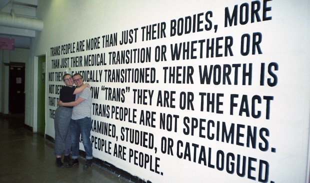 Photo of the two artists Kat and Samuel hugging in front of their mural/installation art piece, black text written on a white wall.