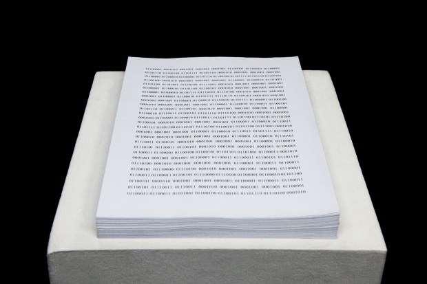 Photo of a text installation featuring binary code printed onto a stack of letter size paper. 