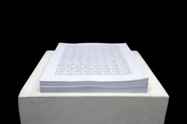 Photo of a text installation featuring binary code printed onto a stack of letter size paper. 