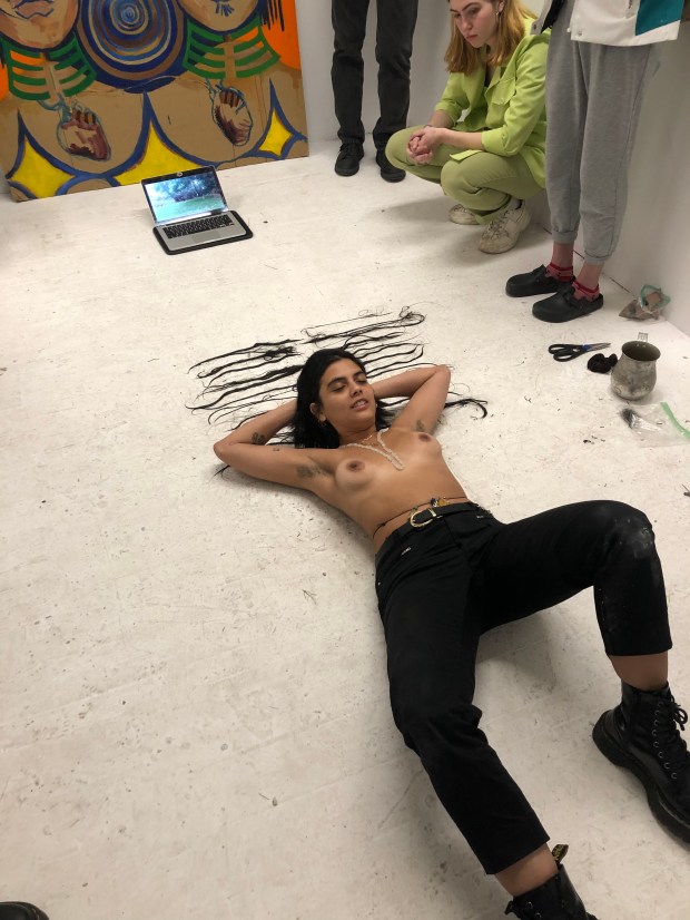 Photo of the artist laying on the ground, topless with tan lines, wearing black jeans, with head resting on strands of hair.