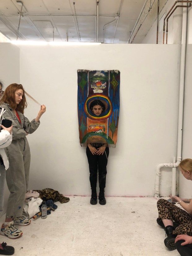 Photo of the artist standing in front of a white wall with head poking through a coloured painting.
