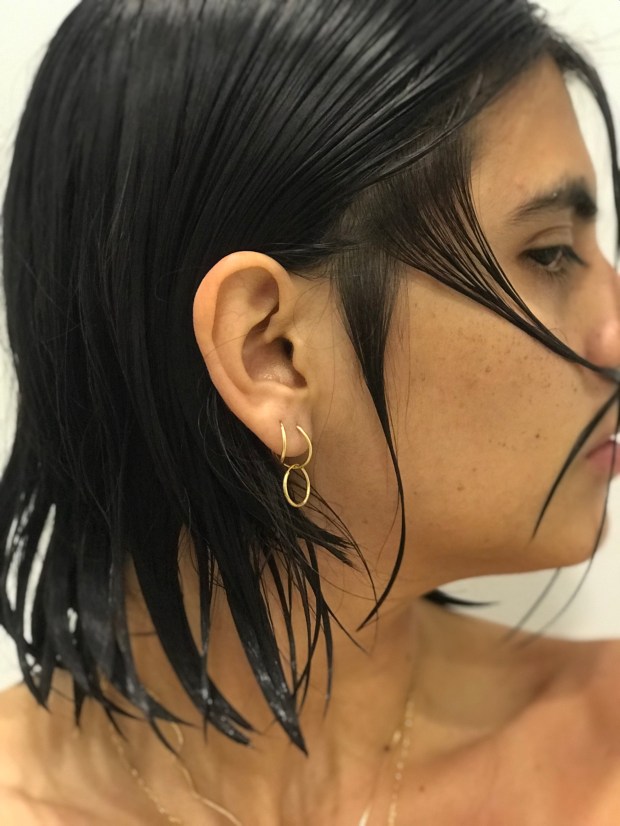 Photo of the artist with slicked shoulder length hair and bare shoulders, with face to the side of the camera. The slicked hair is brushed in strips across the artist’s upper lip like a moustache.