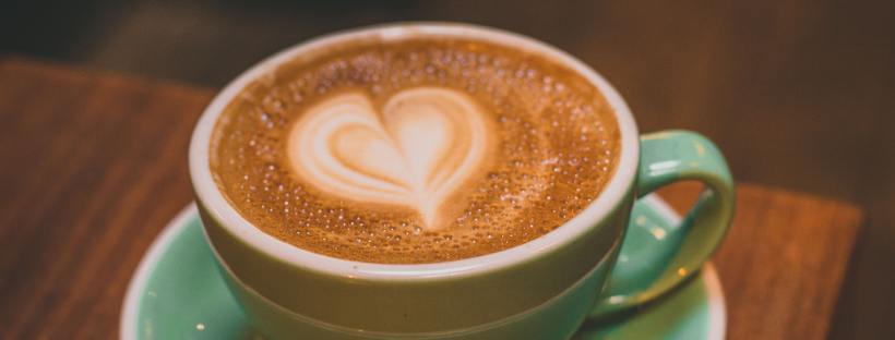 Photo of a green coffee cup and saucer sitting on a brown table, the top of the coffee foam is in the shape of a heart