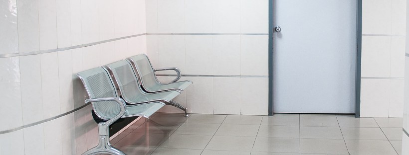 Photo of a white washed waiting room with plastic chairs, a door and a painting on the wall