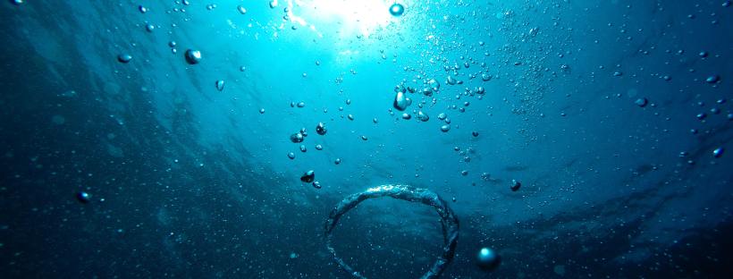 Underwater photo of blue sea with bubbles and a bright light shining above the surface.
