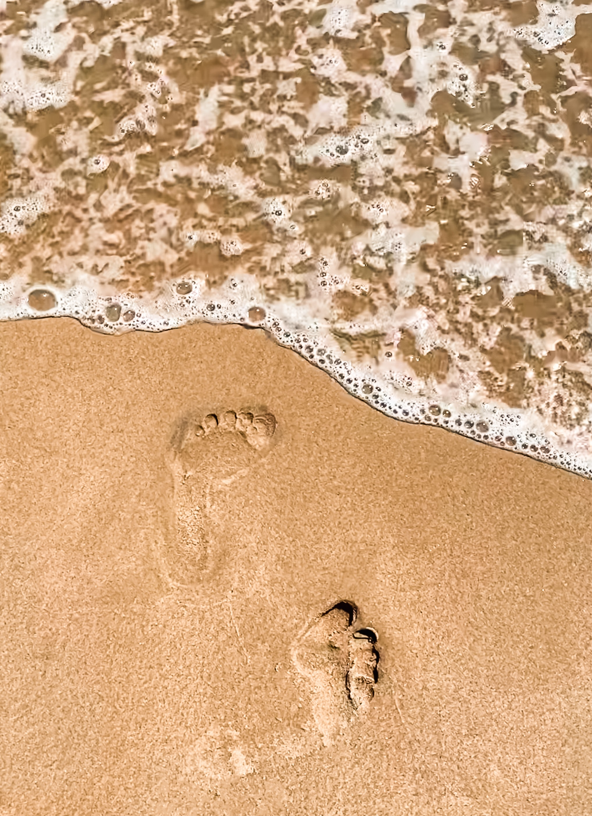 Photo of sea foam on the beach with two footprints in the sand.