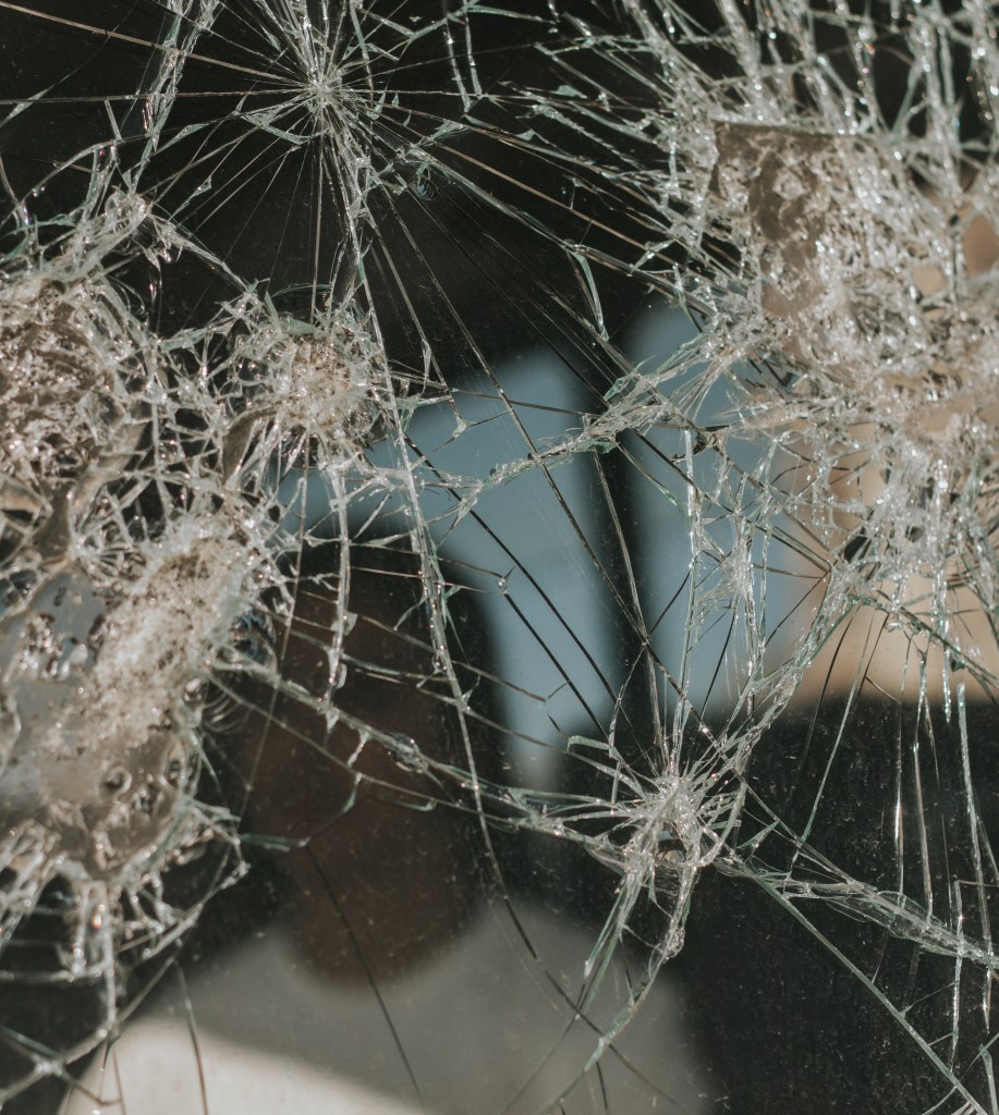 Photo of a broken mirror with figure of a person in a white t-shirt in the reflection.