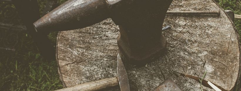 Photo of a metal anvil on a cut log with hammer and tools, surrounded by green grass.