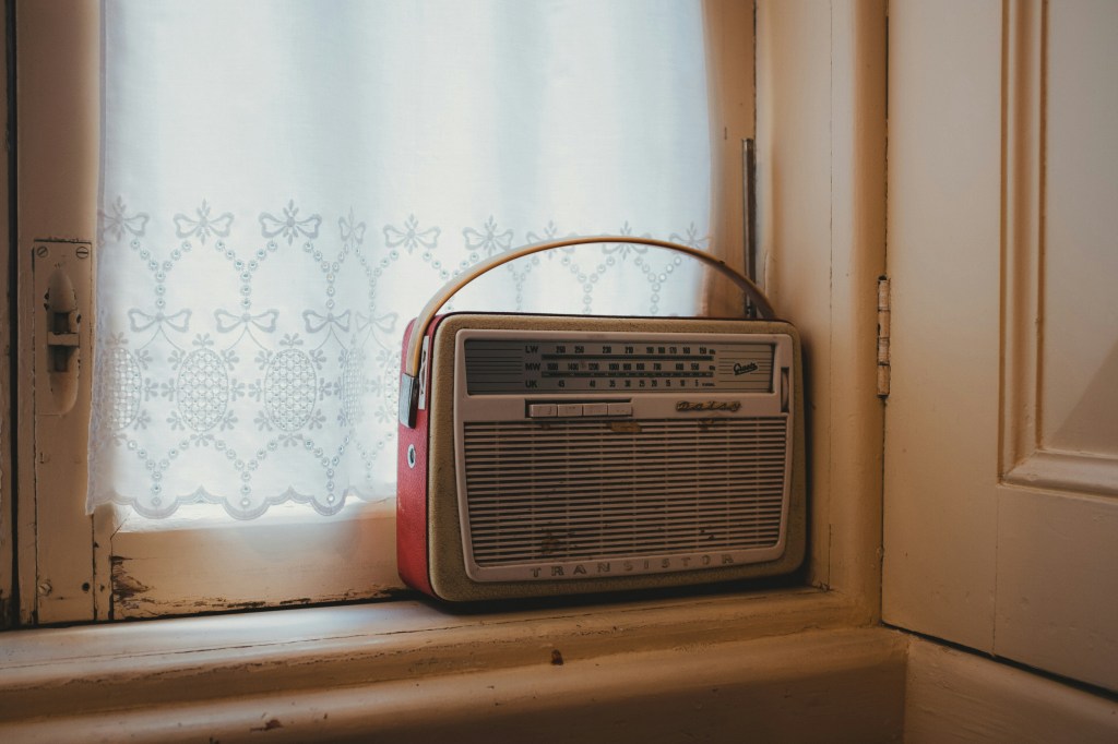 A vintage red and cream-coloured transistor radio sits on a windowsill in front of a white lace curtain with a floral and bow pattern.
