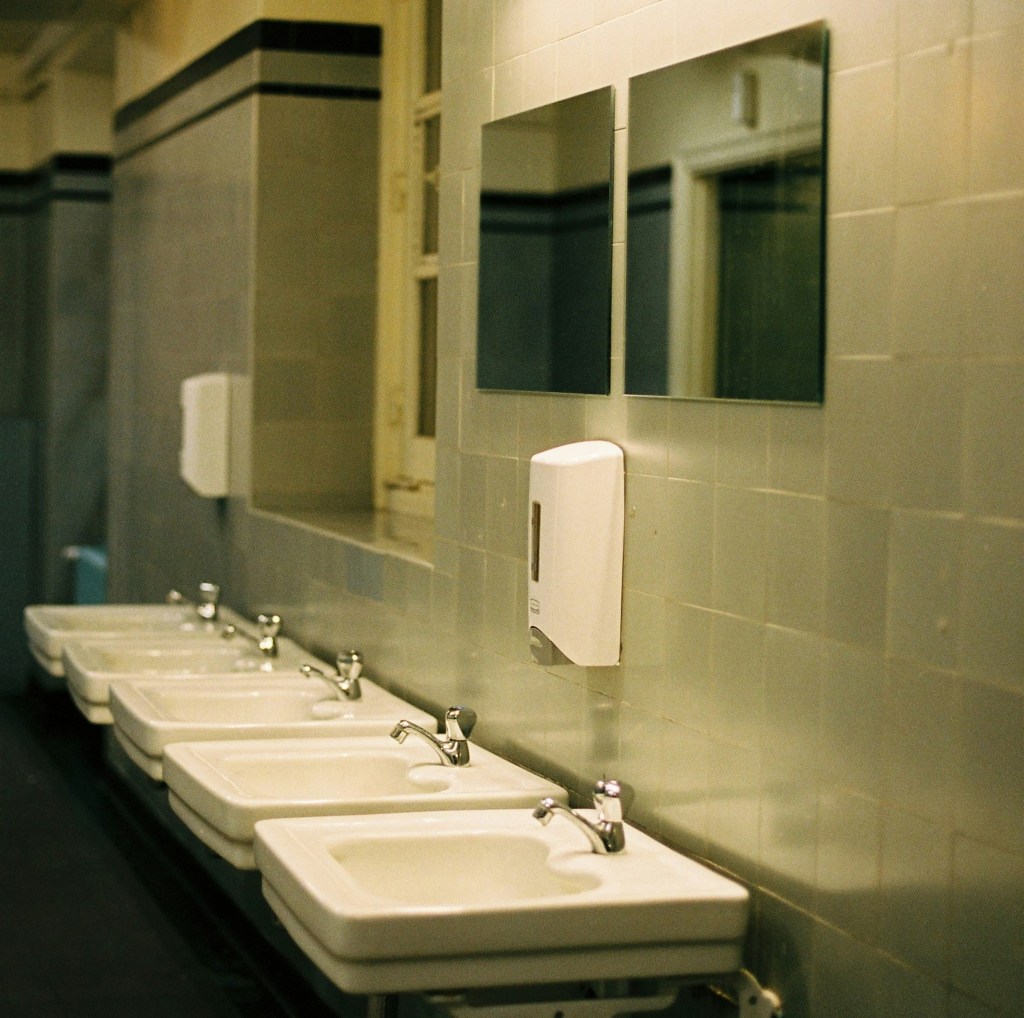 Photo of a public bathroom with beige and white colours and dim lighting, including several mirrors, sinks and handwash dispensers. 