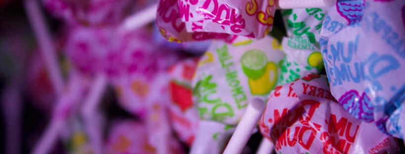 A close-up Photo of a pile of colourful Dum Dums lollipops, each wrapped in bright, patterned wrappers. The lollipops are packed closely together, with white sticks protruding in different directions.