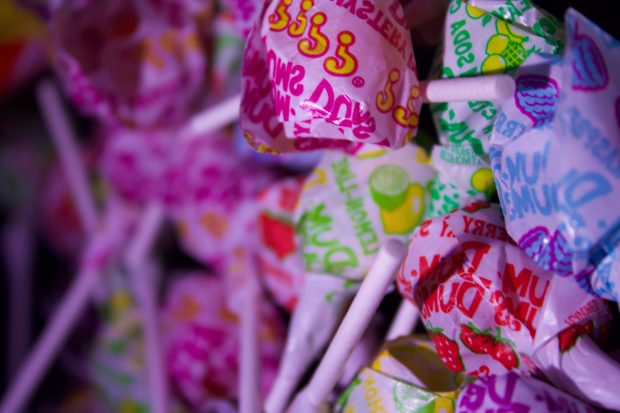 A close-up Photo of a pile of colourful Dum Dums lollipops, each wrapped in bright, patterned wrappers. The lollipops are packed closely together, with white sticks protruding in different directions.