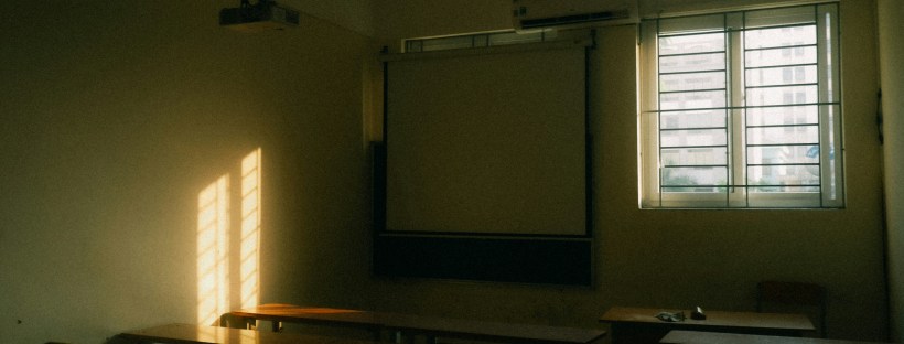 Photo of a darkened classroom with chairs and a whiteboard. A window with open blinds creates some light in the room.