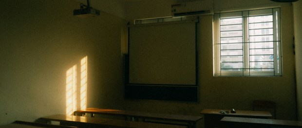 Photo of a darkened classroom with chairs and a whiteboard. A window with open blinds creates some light in the room. 