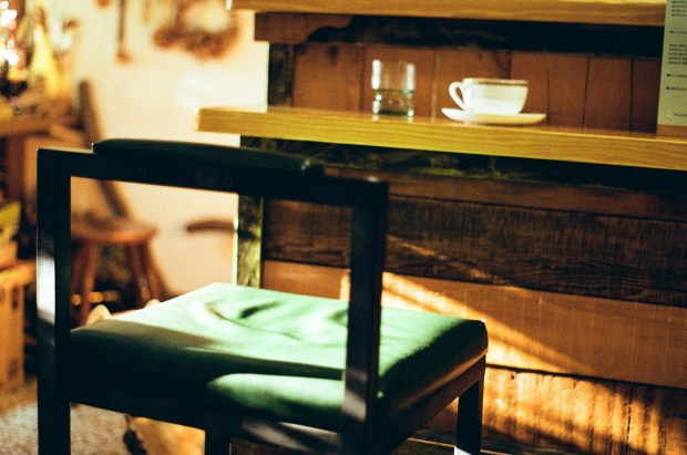 Photo of a wooden stool in a cafe with a green cushion, an empty water glass and coffee cup on the bar table in front.
