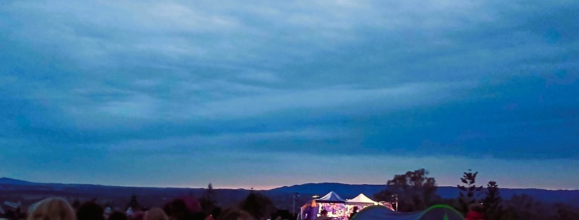 Photo of a campsite at dawn, with people lounging around on grass, with tents and trees in the distance, and a light blue cloudy sky above them.