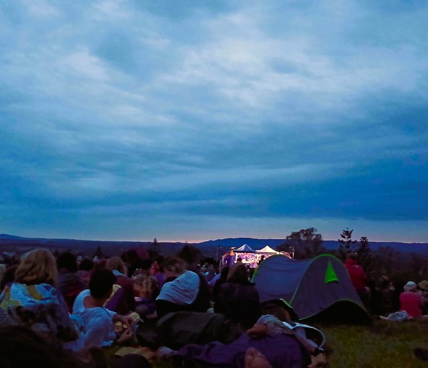 Photo of a campsite at dawn, with people lounging around on grass, with tents and trees in the distance, and a light blue cloudy sky above them.