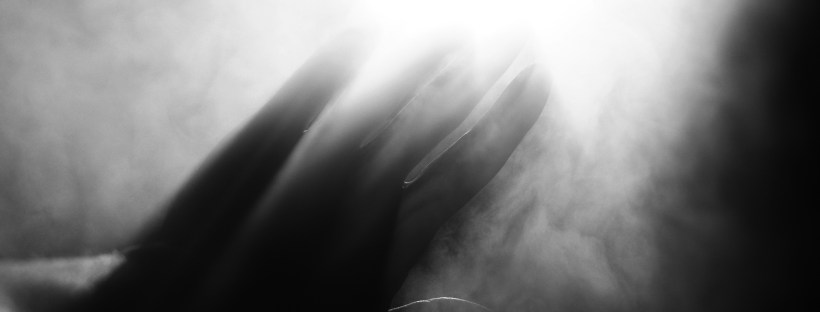Black and white photo of a hand reaching towards bright light through smoke.