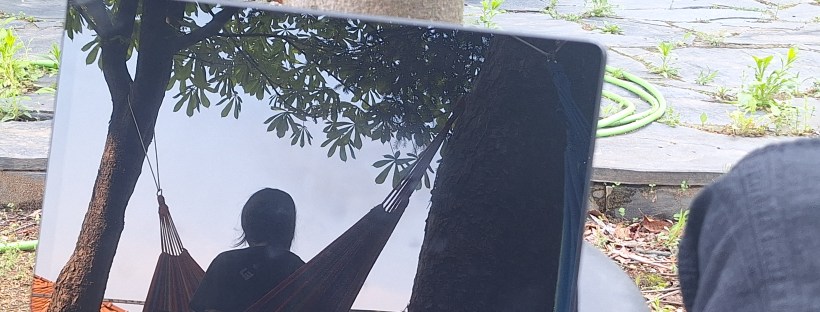 Photo of a person sitting in a hammock reflected in a mirror. The mirror is placed on a stool in the foreground, and the rest of the photo shows a paved garden with a brick wall and fence.