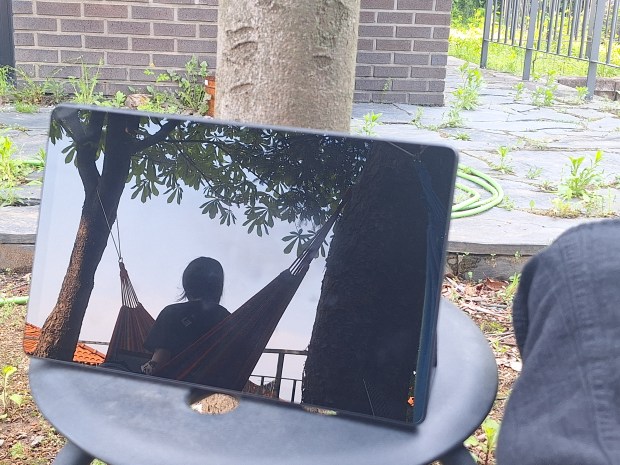 Photo of a person sitting in a hammock reflected in a mirror. The mirror is placed on a stool in the foreground, and the rest of the photo shows a paved garden with a brick wall and fence.