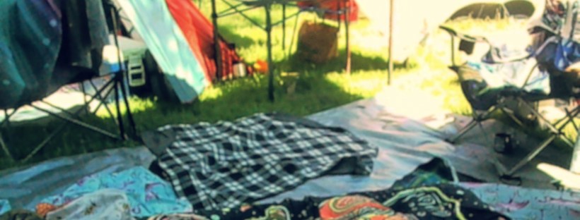Photo of a colourful sunlit campsite with patterned fabric strewn on the grass, and tents and camp chairs in the background.