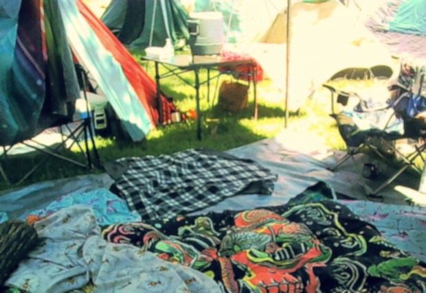 Photo of a colourful sunlit campsite with patterned fabric strewn on the grass, and tents and camp chairs in the background. 