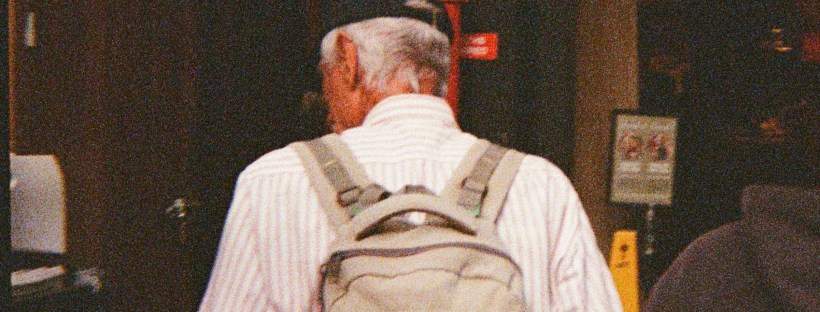 A grainy photo of the back of a person, who has greying hair, a black cap, a white shirt and is carrying a beige backpack.
