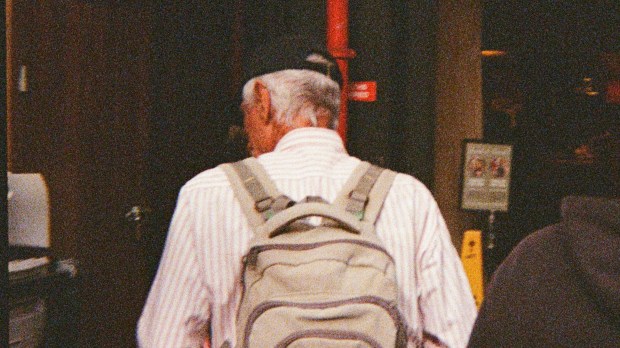 A grainy photo of the back of a person, who has greying hair, a black cap, a white shirt and is carrying a beige backpack.