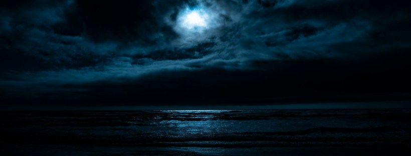 Photo of the sea at night, with dark waves, cloudy sky and a glowing moon.