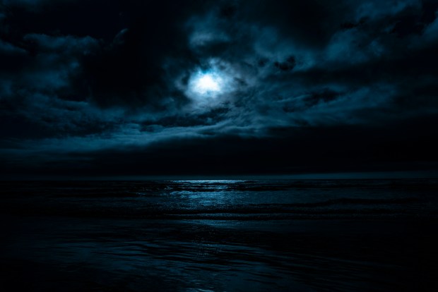 Photo of the sea at night, with dark waves, cloudy sky and a glowing moon.