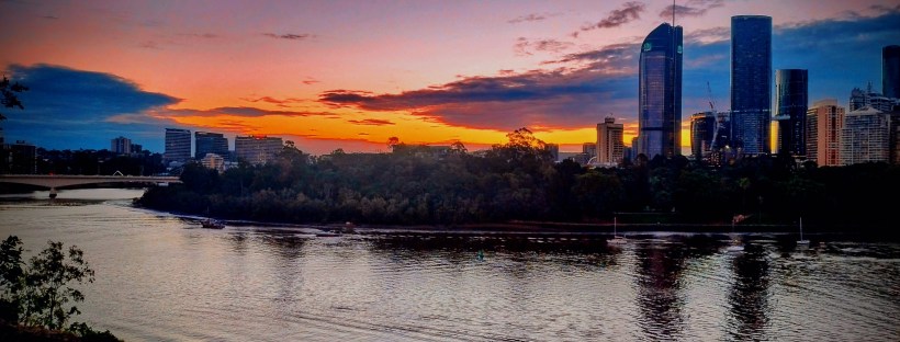Photo taken from a rocky riverside with straggly green plants. The photo shows us the rippling Magandjin river, tall trees and high rises on the opposite side, and an orange and blue sunset with smatterings of clouds.