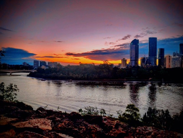 Photo taken from a rocky riverside with straggly green plants. The photo shows us the rippling Magandjin river, tall trees and high rises on the opposite side, and an orange and blue sunset with smatterings of clouds. 