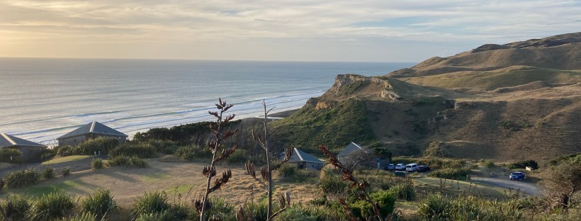 Photo overlooking a valley of hills, green foliage, houses and cars, with a blue sea and sky on the horizon.