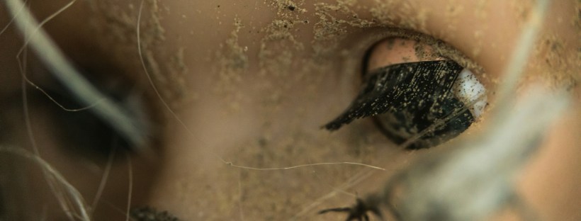 Close up photo of a toy doll’s face, covered in dirt and sand, with their eye half open.