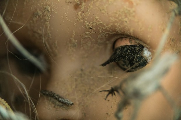 Close up photo of a toy doll’s face, covered in dirt and sand, with their eye half open.
